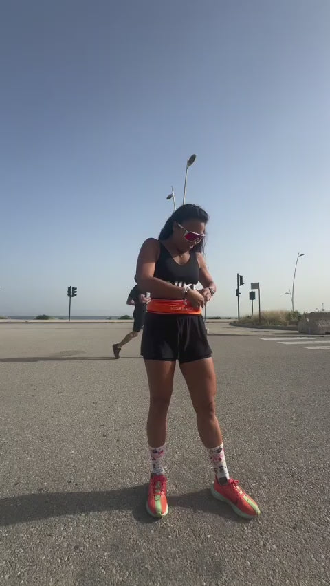 Video of a runner wearing an orange Vitaract running belt jogging along a coastal road, demonstrating the belt staying in place with no bouncing during a real run