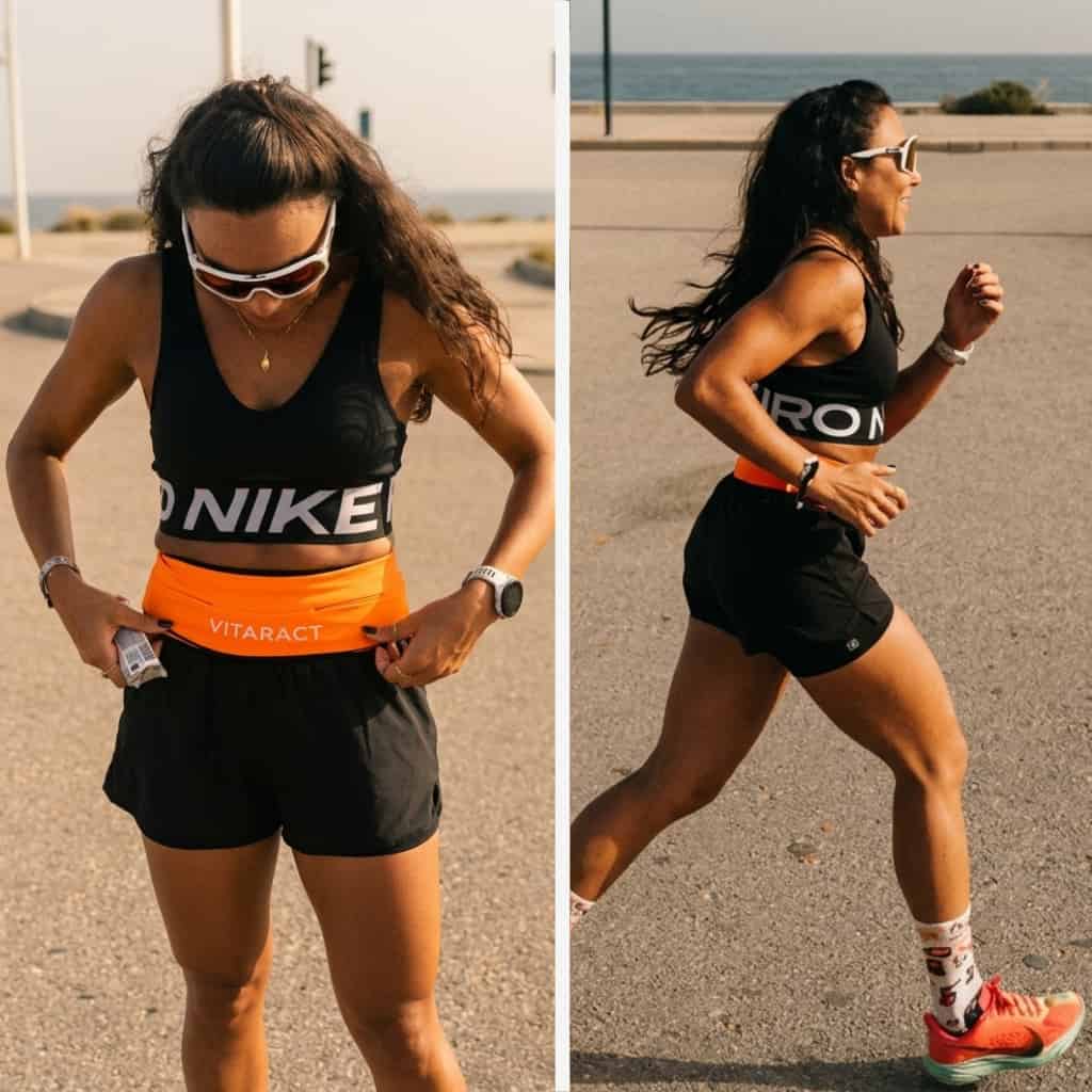 Woman wearing an orange Vitaract running belt while running outdoors near the coast — shown adjusting the belt and running in profile, demonstrating a secure bounce-free fit